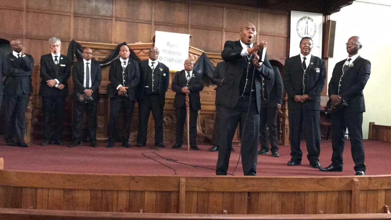 Image of 11 Men in URCSA CMM uniform - a white shirt, black suit and black long pants. 
										The mend are standing in front of the church podium that is decorated with a cloth written 
										'Ruri Moren o tsohile'. One of the men is standing in front of other and is talking on a microphone.