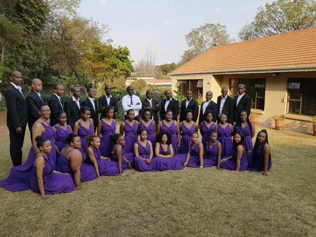 Melodi ya Tshwane choir members posing at a garden in three rows. On the first row 
										are women wearing purple dresses sitting on the grass of the garden. On the second 
										row are women kneeling on the garden grass wearing purple dresses. On the third row 
										are men wearing black suits and white shirts, standing. On the background on the left 
										there is a townhouse and on the right there are trees.