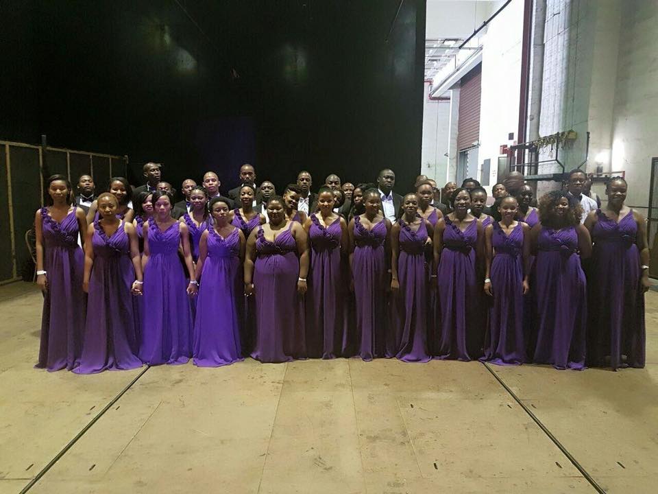 Melodi ya Tshwane choir members posing at a backstage in three rows. On the first and 
										second rows are women wearing purple dresses standing. On the third row are men wearing 
										black suits and white shirts also standing.